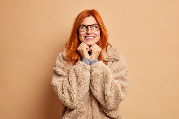 Portrait of good looking redhead woman keeps hands under chin looks away happily dreams about something dressed in brown coat.