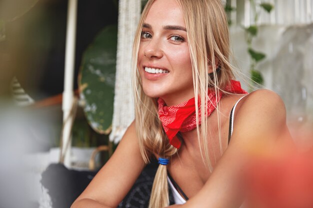 Portrait of good looking relaxed young female model with red bandana on neck, has her own style, sits against cafe cozy interior