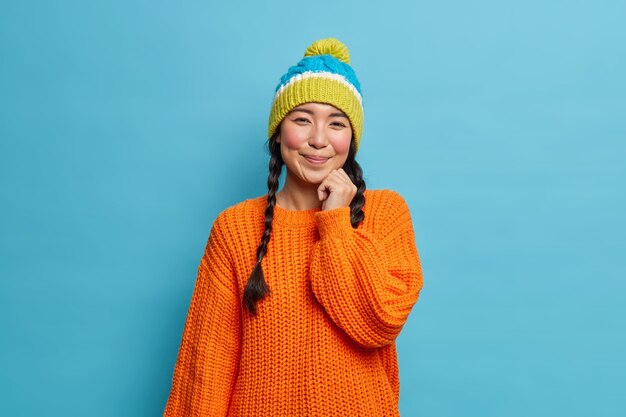 Portrait of good looking shy brunette Asian woman with two pigtails wears knitted sweater and hat has satisfied face expression poses in studio against blue wall walks during winter time