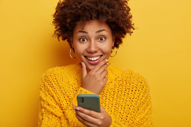 Portrait of good looking smiling female touches chin, smiles broadly, uses cellphone, wears yellow sweater, models indoor.