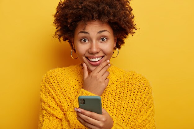 Portrait of good looking smiling female touches chin, smiles broadly, uses cellphone, wears yellow sweater, models indoor.