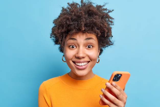 Free photo portrait of good looking smiling young woman with curly hair uses mobile phone for chatting online downloads new app looks gladfully  wears orange jumper isolated over blue wall