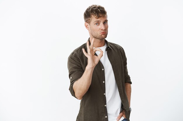 Portrait of good-looking sportsman pulling lips up and showing okay gesture being impressed by good skills or product standing satisfied and affected over gray wall