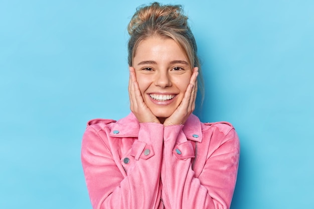 Portrait of good looking woman smiles positively keeps hands on cheeks has healthy skim white teeth hears something pleasant wears pink jacket isolated over bluebackground. Happy emotions concept