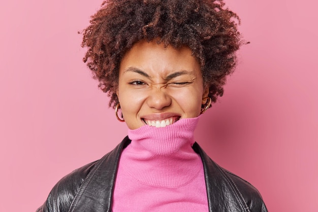 Foto gratuita il ritratto di bella donna con i capelli ricci strizza l'occhio stringe i denti copre il mento con il colletto a collo alto vestito con un'elegante giacca di pelle ha un'espressione giocosa isolata su sfondo rosa