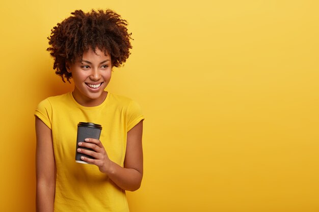 Portrait of good looking woman with curly hairstyle, has thoughtful satisfied facial expression, enjoys hot aromatic coffee, wears yellow t shirt