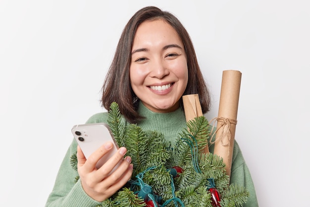 Portrait of good looking woman with dark hair smiles broadly holds mobile phone messages with friends in chat holds spruce branches and paper rollers isolated over white backgroud. Winter time