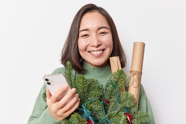 Portrait of good looking woman with dark hair smiles broadly holds mobile phone messages with friends in chat holds spruce branches and paper rollers isolated over white backgroud. Winter time