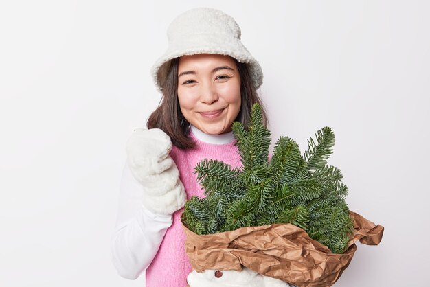 Portrait of good looking young Asian woman smiles gently wears warm winter panama and mittens holds bouquet of spruce branches going to make New Year decoration isolated over white background
