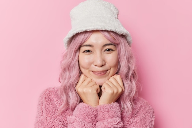 Free photo portrait of good looking young asian woman with dyed hair keeps hands under chin smiles gently looks directly at camera weas stylish hat and winter coat poses against pink background in studio.