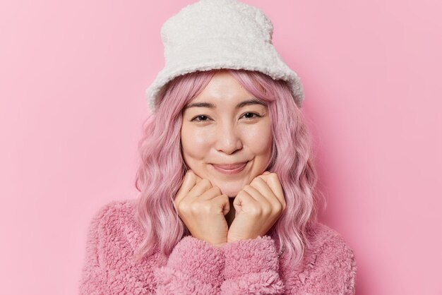 염색된 머리를 한 잘 생긴 젊은 아시아 여성의 초상화는 턱 미소 아래 손을 유지하고 스튜디오의 분홍색 배경에 대해 세련된 모자와 겨울 코트 포즈를 취하는 카메라를 부드럽게 직접 쳐다봅니다.