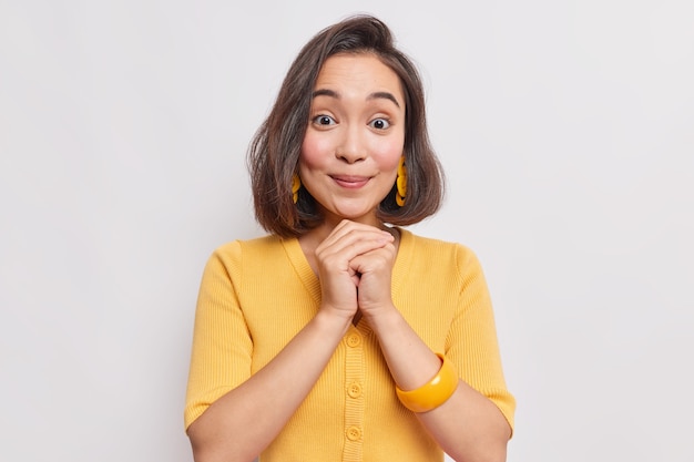 Portrait of good looking young Asian woman with natural dak hair healthy skin keeps hands together under chin wears yellow earrings jumper and bracelet on arm isolated over white wall