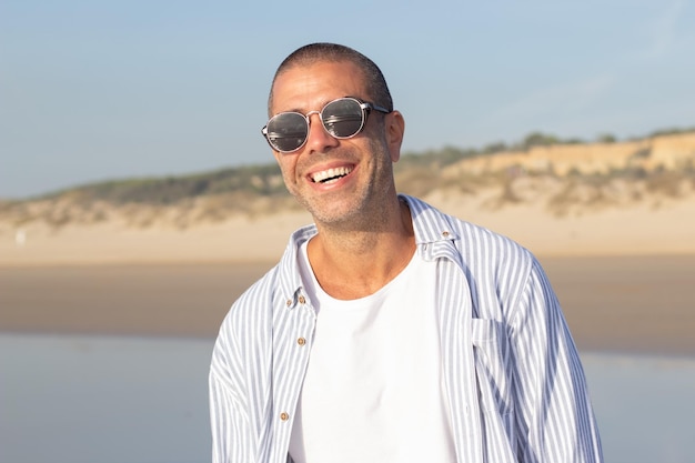 Portrait of good-looking young man on beach. Male model with shaved head in sunglasses smiling brightly at camera. Portrait, vacation, beauty concept