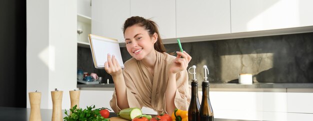 ノートを持ち、レシピ料理を読む食料品リストを持つ格好良い笑顔の女の子の肖像画