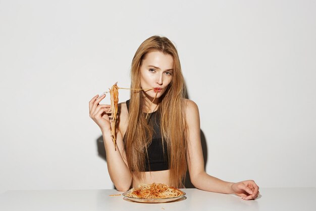 Портрет красивой молодой женщины с длинными светлыми волосами в черном топе, поедающей спагетти вилкой и смотрящей в камеру с расслабленным взглядом
