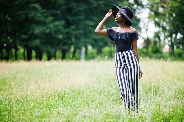검은색과 흰색 줄무늬 바지를 입고 공원의 푸른 잔디에서 포즈를 취한 여름 모자를 쓴 20대 멋진 아프리카계 미국인 여성의 초상화