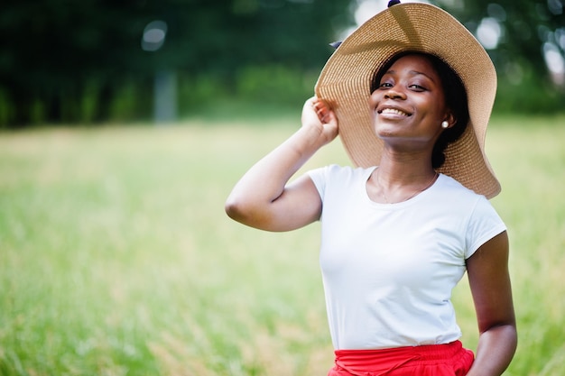 Портрет великолепной африканской американки 20 лет в летней шляпе, красных штанах и белой футболке, позирующей на зеленой траве в парке