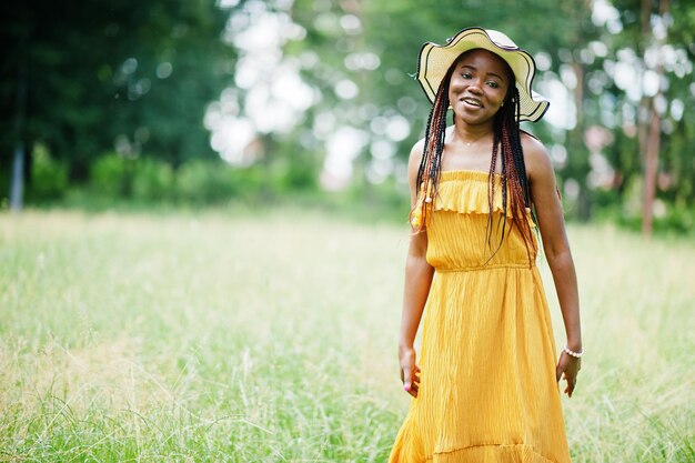 公園の緑の芝生でポーズをとって黄色のドレスと夏の帽子を身に着けているゴージャスなアフリカ系アメリカ人女性20代の肖像画
