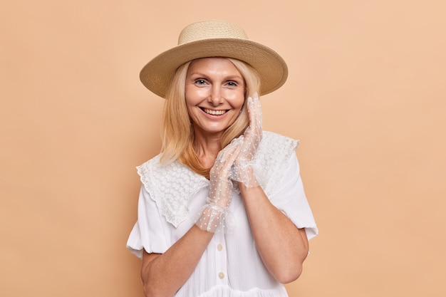 Portrait of gorgeous aristocratic woman with pleasant appearance smiles happily keeps hands together wears fedora white dress and lace gloves expresses positive emotions poses against beige wall