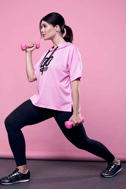 Портрет шикарной фигуры позитивной латинской женщины в розовой спортивной толстовке с гантелями на розовом