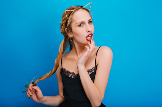 Portrait of gorgeous girl with sexy look, touching her red lips at party. She has long blonde hair, nice manicure. Wearing black dress, diadem with cat ears.