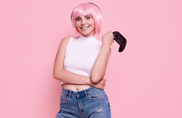 Portrait of gorgeous happy gamer girl with pink hair playing video games using joystick on colorful in studio