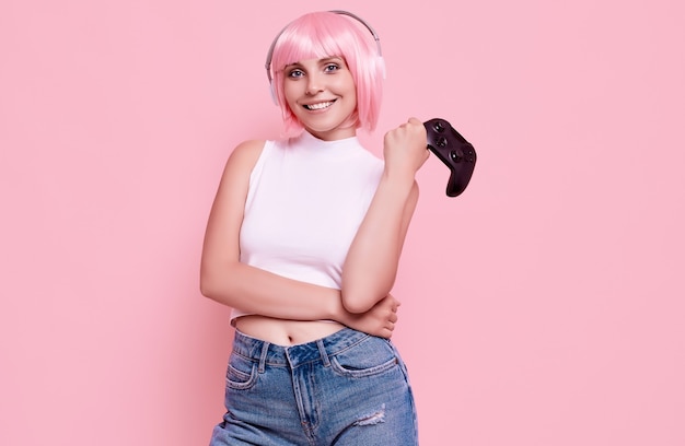 Portrait of gorgeous happy gamer girl with pink hair playing video games using joystick on colorful in studio