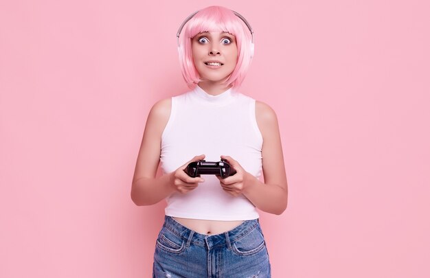 Portrait of gorgeous happy gamer girl with pink hair playing video games using joystick on colorful in studio