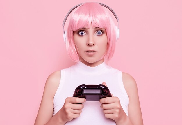 Portrait of gorgeous happy gamer girl with pink hair playing video games using joystick on colorful in studio