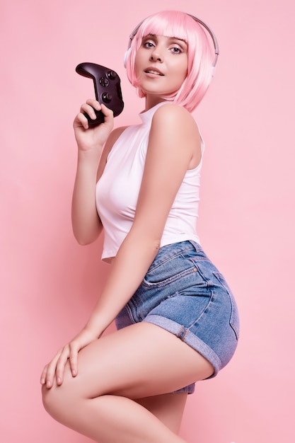 Portrait of gorgeous happy gamer girl with pink hair playing video games using joystick on colorful in studio
