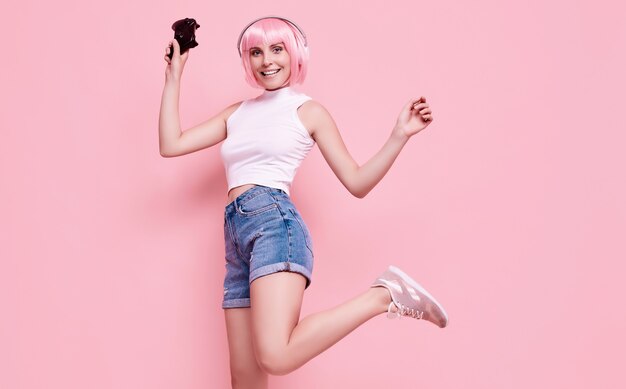 Portrait of gorgeous happy gamer girl with pink hair playing video games using joystick on colorful in studio