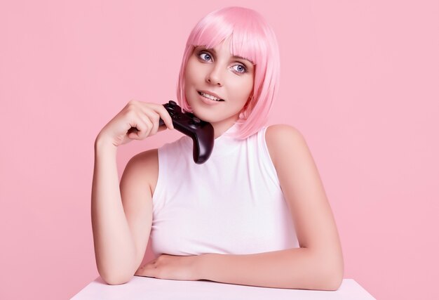 Portrait of gorgeous happy gamer girl with pink hair playing video games using joystick on colorful in studio