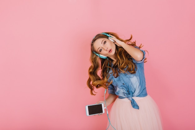 Portrait of gorgeous happy long-haired girl in headphones singing favorite song isolated on pink background. Attractive curly young woman in denim shirt with smartphone dancing and enjoying music