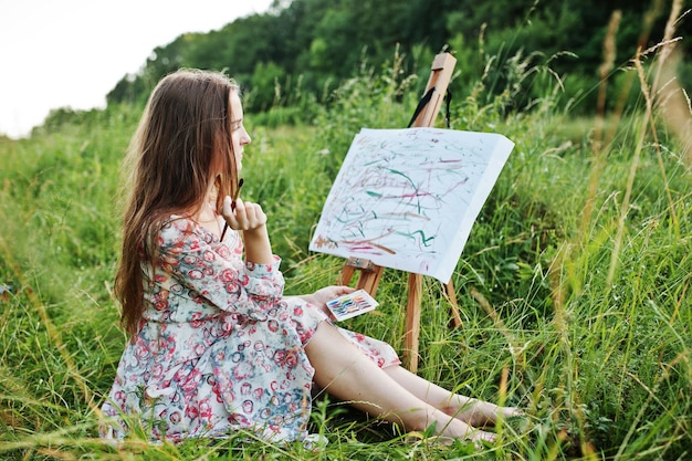 Portrait of a gorgeous happy young woman in beautiful dress sitting on the grass and painting on paper with watercolors