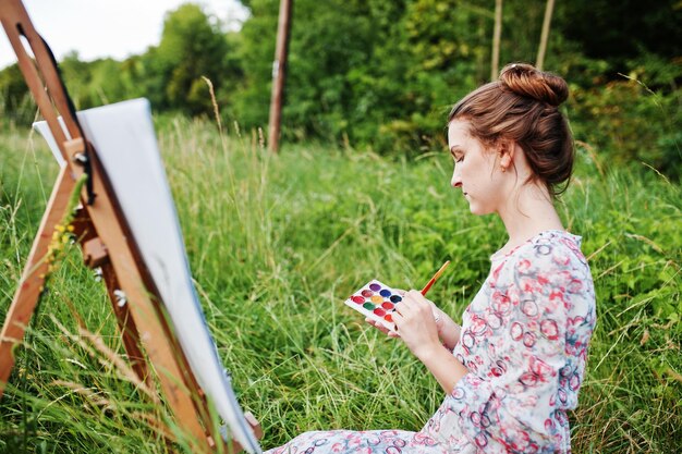 Portrait of a gorgeous happy young woman in beautiful dress sitting on the grass and painting on paper with watercolors