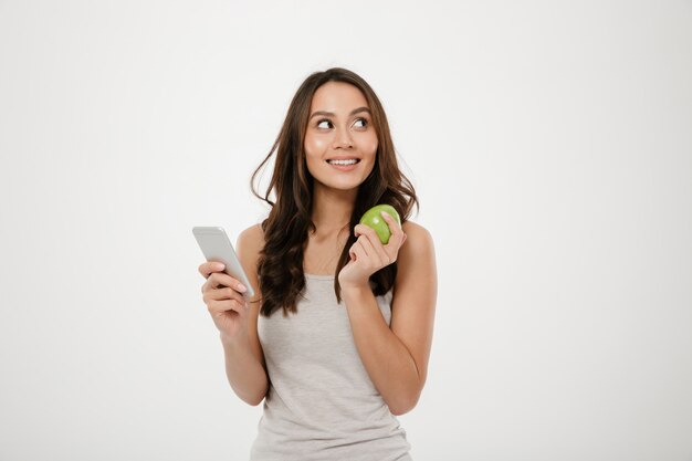 Portrait of gorgeous healthy woman looking aside while posing on camera with green apple and smartphone in hands, isolated over white wall