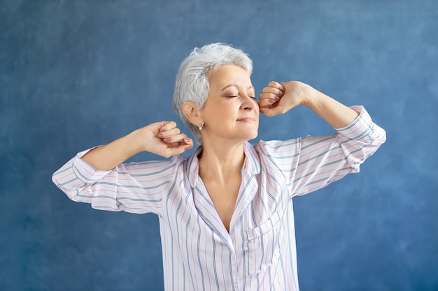 Portrait of gorgeous middle aged woman in stylish striped pajamas stretching body after awakening early in the morning