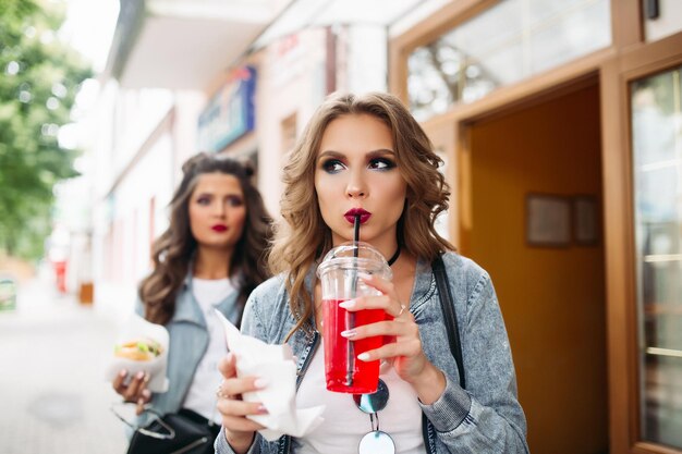 Portrait of gorgeous teenagers with make up and hairstyles drinking lemonade and carrying fast food going out of the cafe.