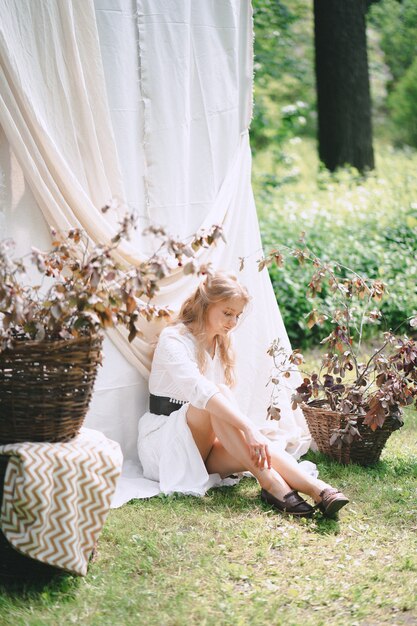 Portrait of gorgeous woman at garden with white wall sitting on grass and thinking in white dress during daytime.