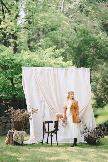 Portrait of gorgeous woman at garden with white wall standing and looking in white dress and brown jacket, during daytime.