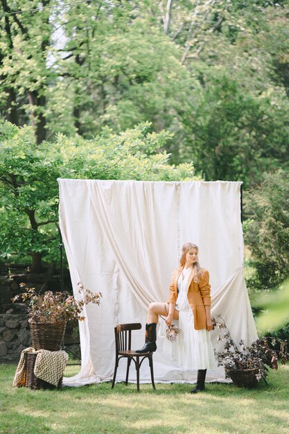 Portrait of gorgeous woman at garden with white wall standing and looking in white dress and brown jacket, during daytime.