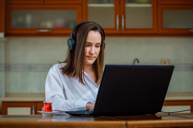 Portrait of gorgeous woman at kitchen working on laptop and with headphones on, in white shirt