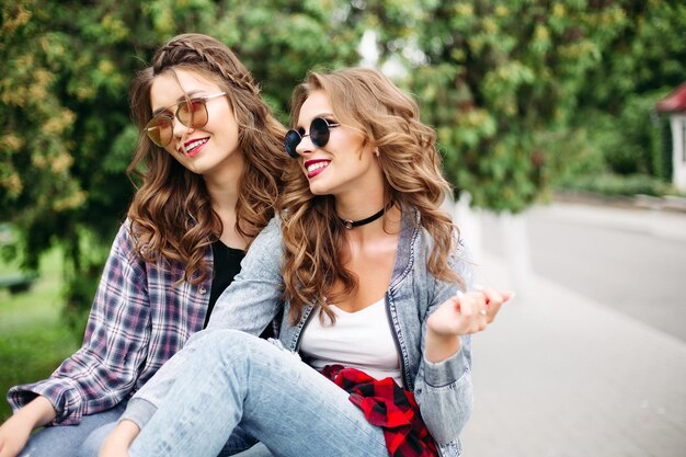 Portrait of gorgeous young girls with brunette and blonde hair in hairstyles wearing fashionable sunglasses and casual clothes smiling at camera in green park at daytime.