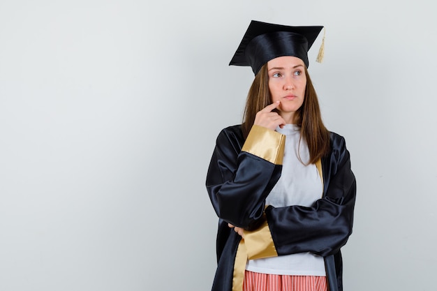 Портрет выпускницы, держащей палец на подбородке в повседневной одежде, униформе и продуманного вида спереди