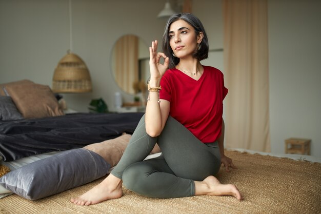 Portrait of gray haired cute young female in casual clothes sitting on floor doing ardha matsyendrasana or sitting half spinal twist, practicing yoga, stimulating digestive system in the morning