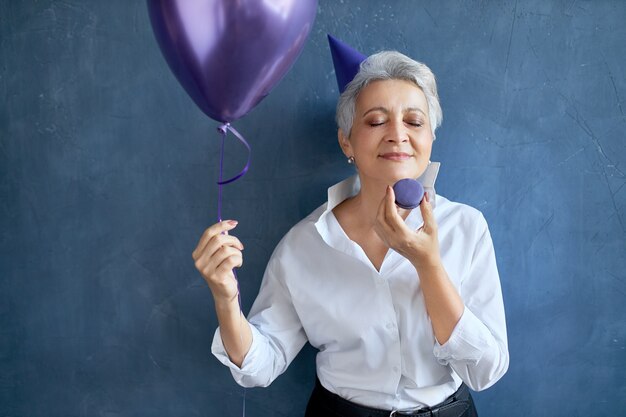 기쁨으로 눈을 감고 생일을 축하하는 세련된 흰색 셔츠에 회색 머리 할머니의 초상화