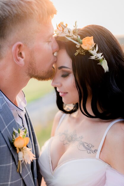 Portrait of groom who is kissing tattooed bride with open decollete and tender wreath made of fresh flowers