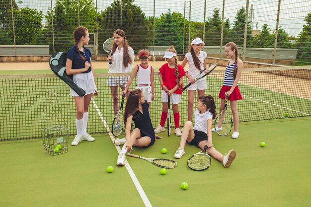 Portrait of group of girls as tennis players holding tennis rackets against green grass of outdoor court. Stylish young teens posing at park. Sport style. Teen and kids fashion concept.