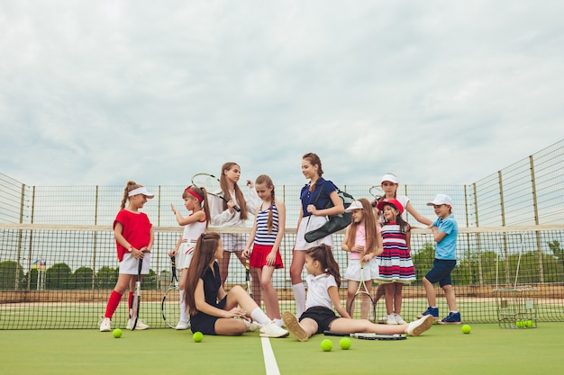 Портрет группы в составе девушки и мальчик как теннисисты держа ракетки тенниса против зеленой травы открытого суда.