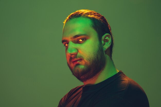 Portrait of a guy with colorful neon light on green  wall. Male model with calm and serious mood. Facial expression, millenials lifestyle and look like. Future, technologies.